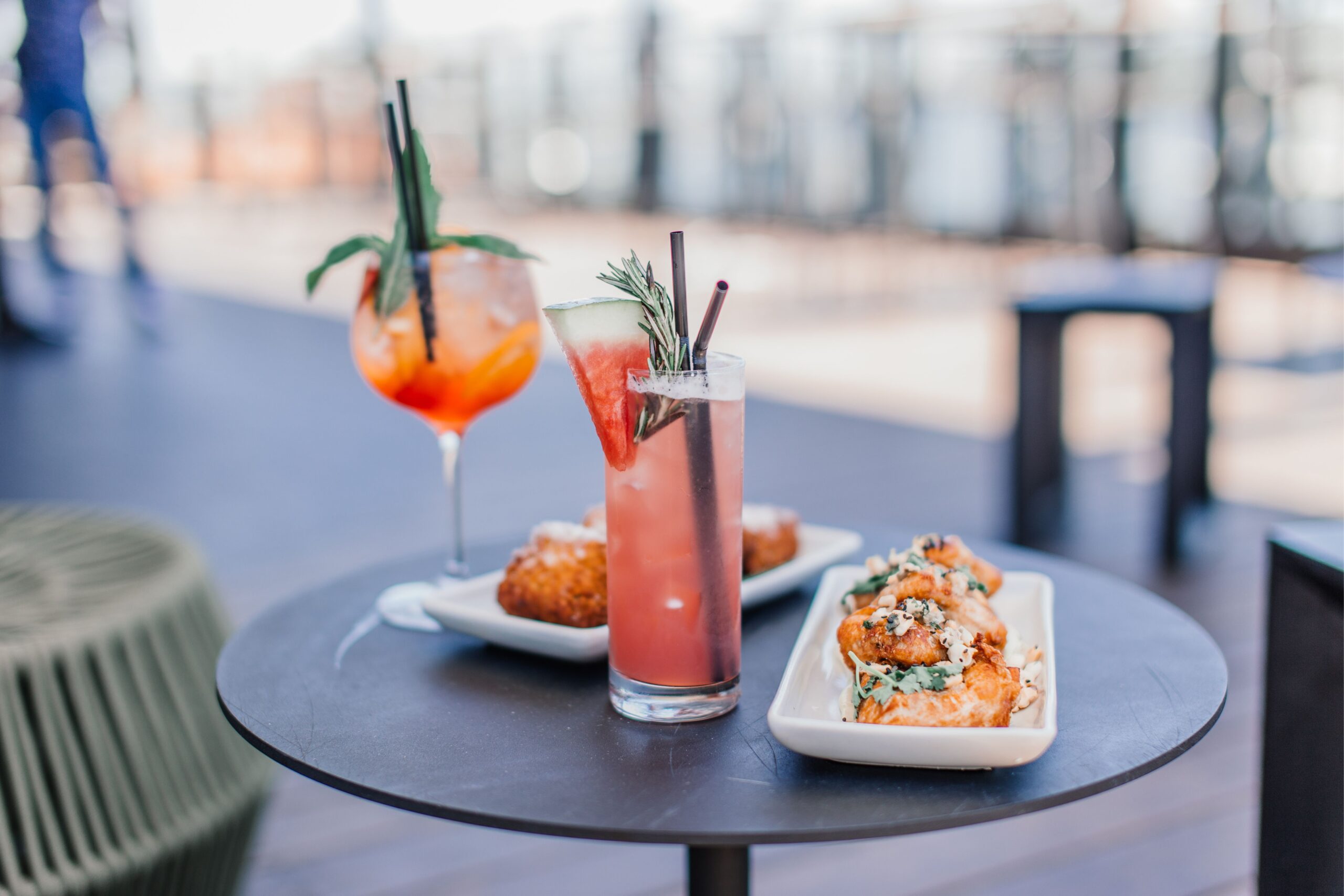 sommerdrinks 2025 Zwei bunte Cocktails und Teller mit frittierten Snacks auf einem runden Tisch, unscharfer Hintergrund im Freien.