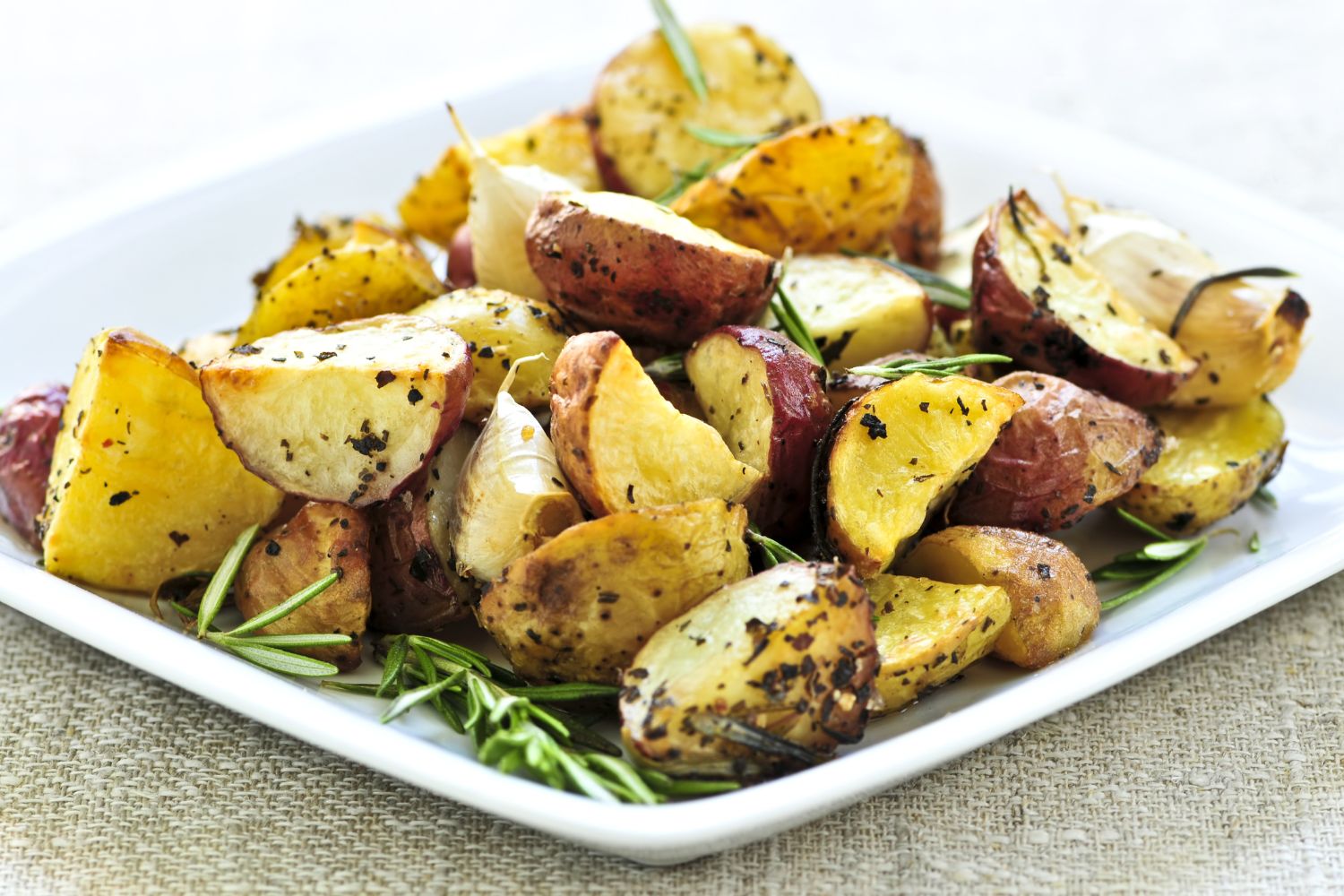 Bratkartoffeln in der Heißluftfritteuse-01 Gebratene Kartoffelstücke mit Kräutern und Rosmarinzweigen auf weißem Teller, auf beigem Tischläufer.