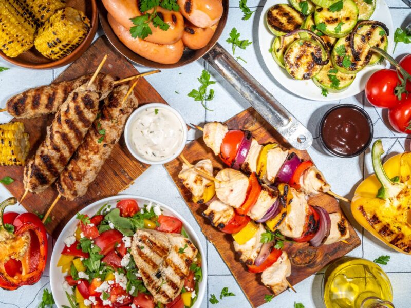 Gegrilltes Gemüse und Fleischspieße, serviert mit Salat, Soßen, Mais und Bratwürsten auf einem gedeckten Tisch.