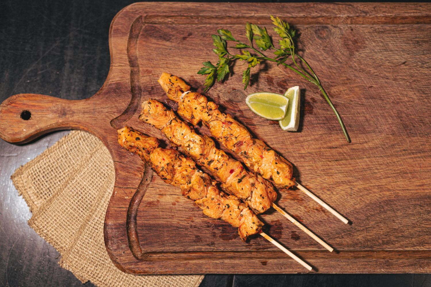 Drei gegrillte Hähnchenspieße auf einem Holzbrett mit Limettenscheiben und Petersilie als Garnierung.