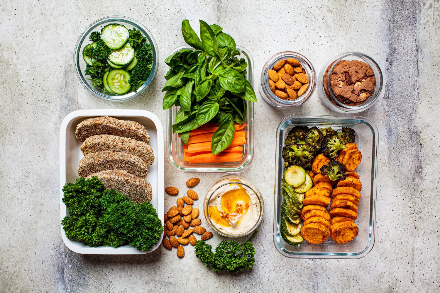 Gesunde Zutaten in Behältern: Gurken, Brokkoli, Karotten, Kräuter, Mandeln, Kekse, Hummus und Brotscheiben arrangiert.