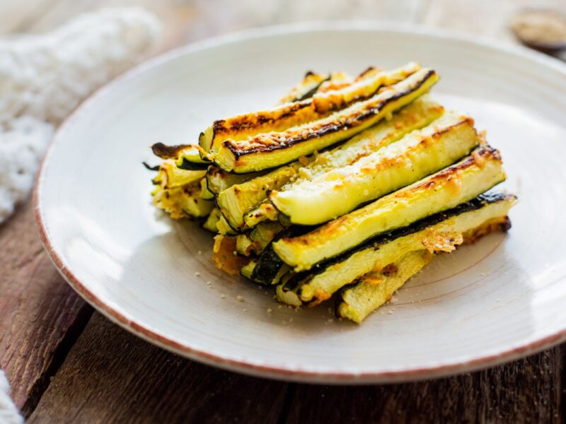 Gebratene Zucchinisticks mit Parmesan auf einem weißen Teller, Holzuntergrund im Hintergrund.