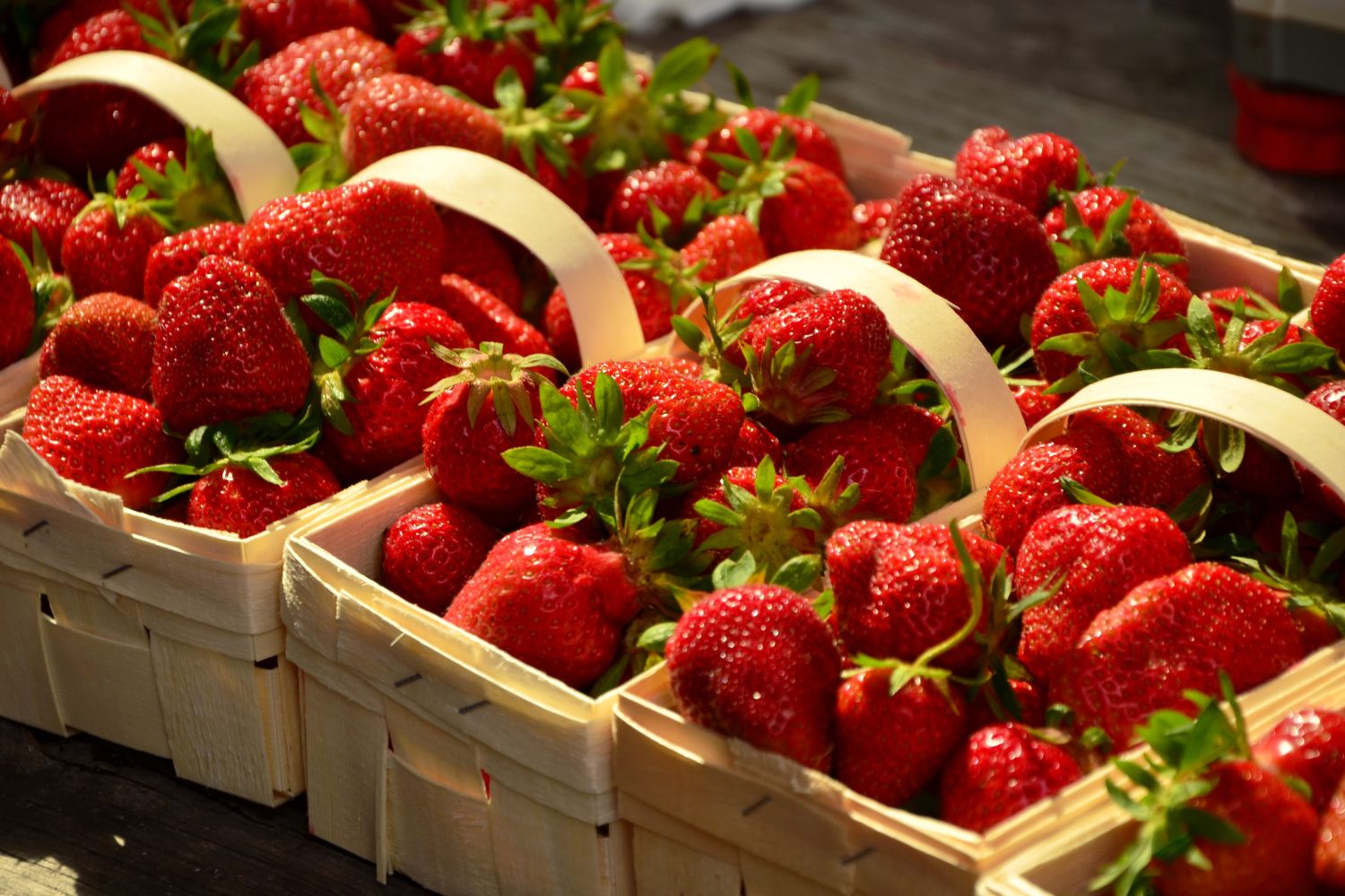 Frische, rote Erdbeeren in Holzschalen mit Griffen auf einem Holztisch, in der Sonne glänzend.