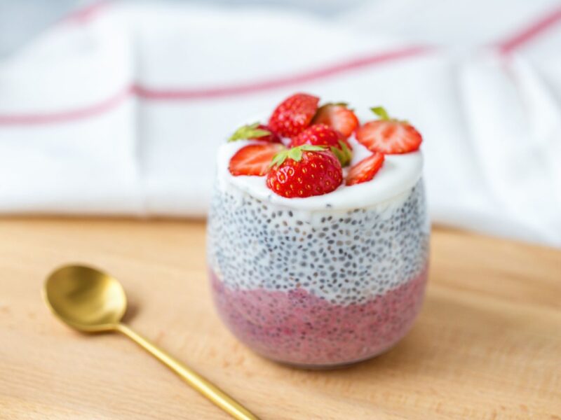 Chia-Pudding im Glas mit Joghurtschicht und frischen Erdbeeren, Goldlöffel daneben auf Holzbrett.