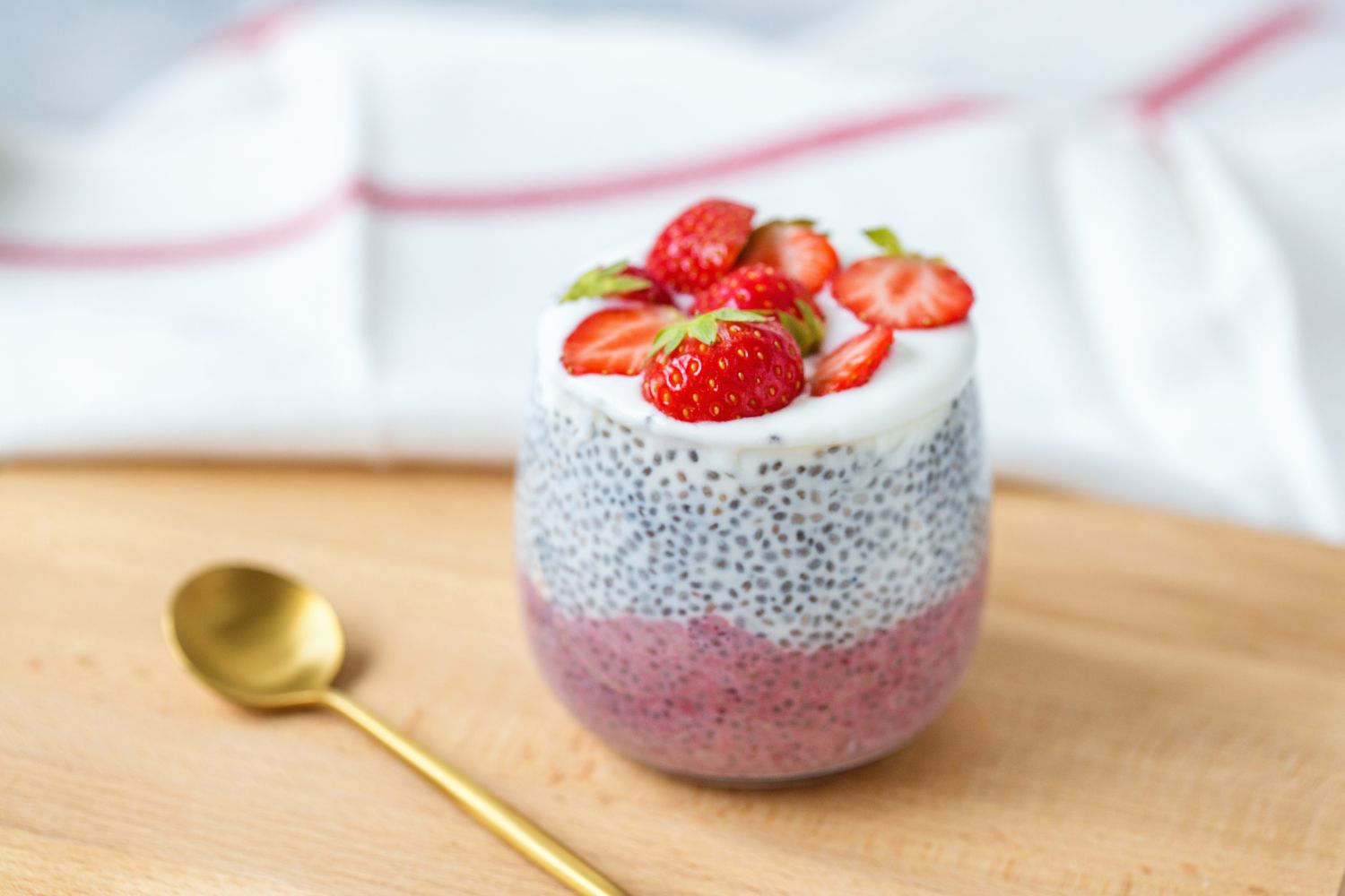 Chia-Pudding im Glas mit Joghurtschicht und frischen Erdbeeren, Goldlöffel daneben auf Holzbrett.