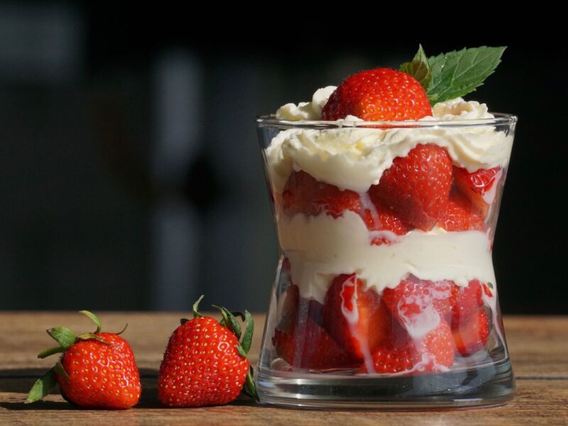 Dessertglas mit Schichten aus Erdbeeren und Sahne, oben mit Erdbeere und Minzblatt, neben drei Erdbeeren auf Holztisch.