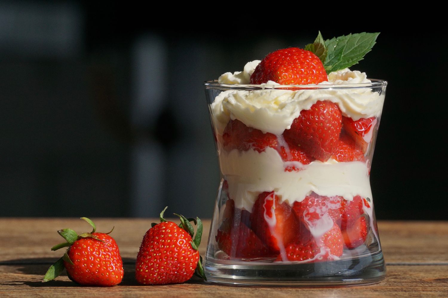 Dessertglas mit Schichten aus Erdbeeren und Sahne, oben mit Erdbeere und Minzblatt, neben drei Erdbeeren auf Holztisch.