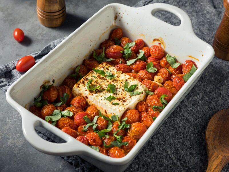 Gebackener Feta mit Kirschtomaten, Knoblauch und Basilikum in einer weißen Auflaufform auf grauem Hintergrund.