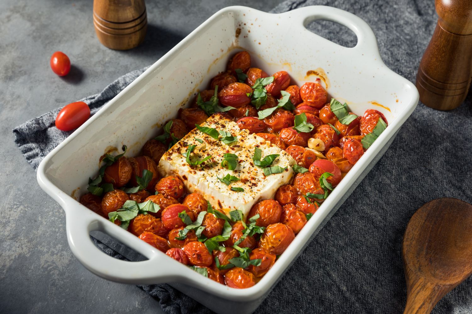 Gebackener Feta mit Kirschtomaten, Knoblauch und Basilikum in einer weißen Auflaufform auf grauem Hintergrund.
