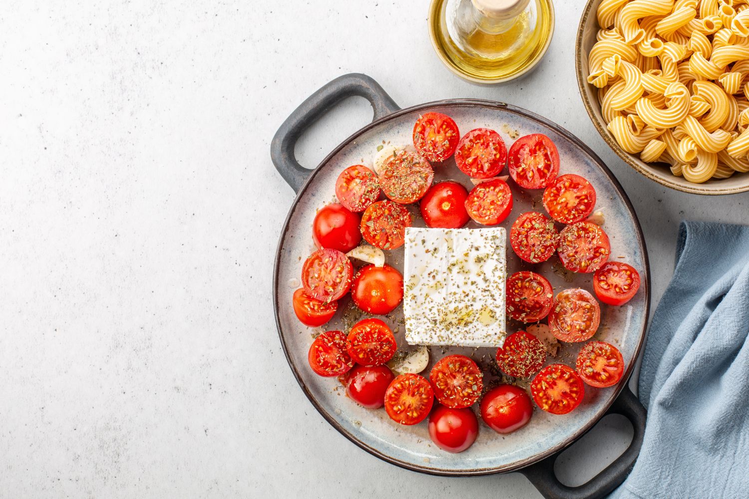 Ofenform mit Tomaten, Feta und Gewürzen. Daneben eine Schale Pasta und eine Flasche Olivenöl auf hellem Hintergrund.
