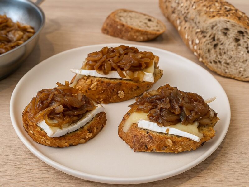 Drei Brotscheiben mit Brie und karamellisierten Zwiebeln auf einem Teller, daneben eine Brotstange und eine Pfanne.