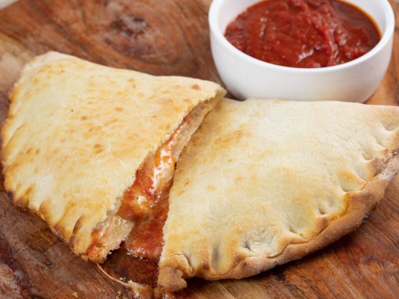 Calzone auf Holzbrett, gefüllt mit Käse und Tomatensauce, daneben ein Schälchen mit weiterer Tomatensauce.