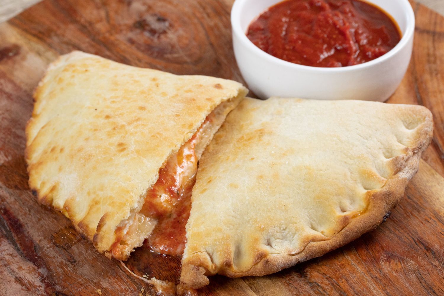 Calzone auf Holzbrett, gefüllt mit Käse und Tomatensauce, daneben ein Schälchen mit weiterer Tomatensauce.