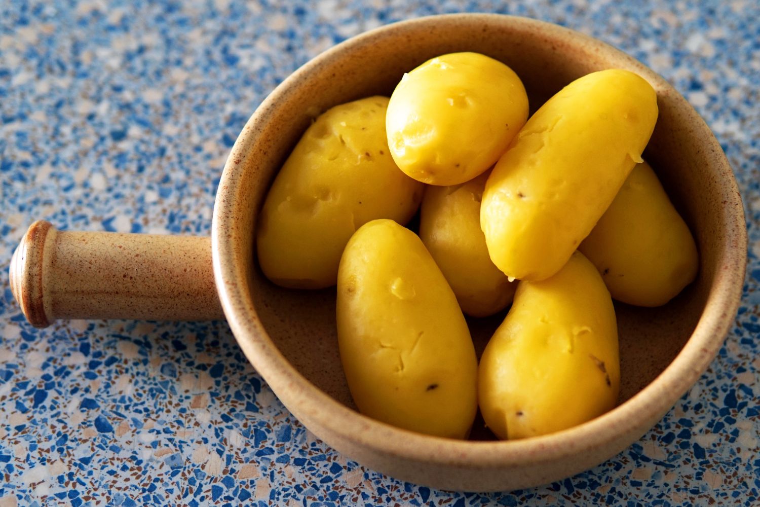 Braune Schale mit gekochten, gelben Kartoffeln auf einer blau gesprenkelten Oberfläche.