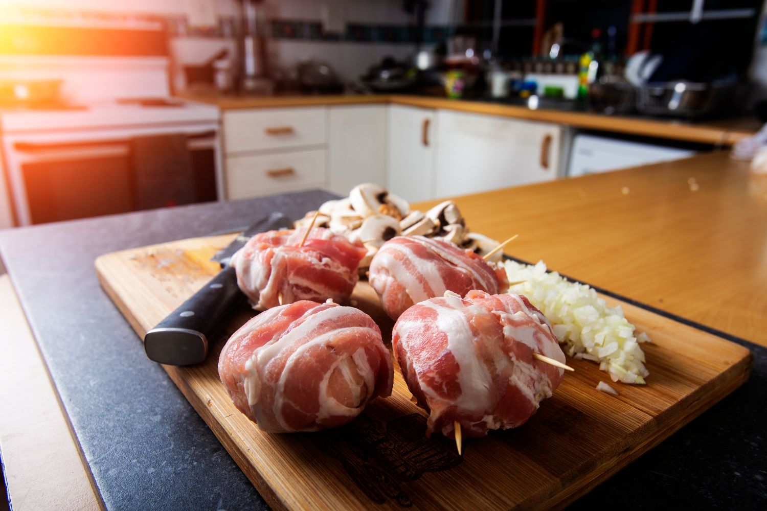 Hackfleischbällchen mit Speck auf Holzbrett, daneben gehackte Zwiebeln und Champignons in einer Küche.
