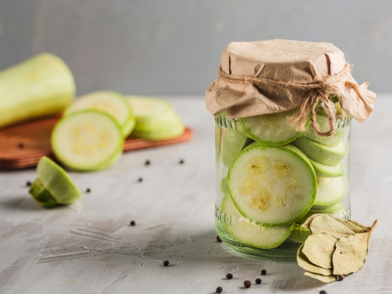 Glas mit eingelegten Zucchinischeiben, Papierabdeckung, Lorbeerblätter und Pfefferkörner auf grauem Untergrund.