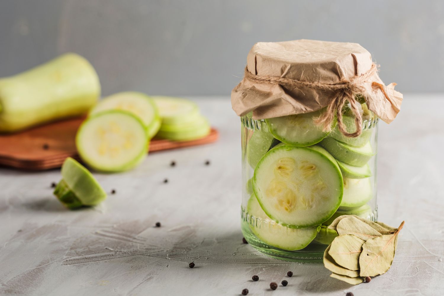 Glas mit eingelegten Zucchinischeiben, Papierabdeckung, Lorbeerblätter und Pfefferkörner auf grauem Untergrund.