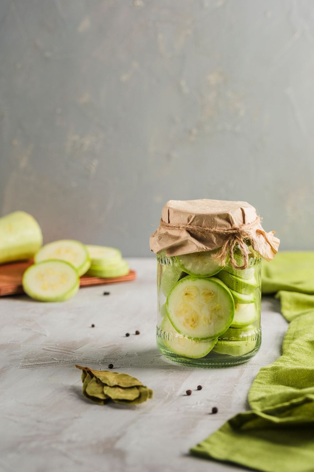 Zucchini Ein Glas mit eingelegten Zucchinischeiben, abgedeckt mit Papier, daneben Zucchinischeiben und Lorbeerblätter.