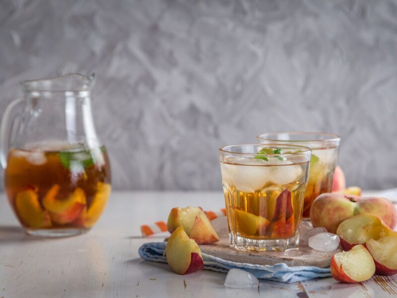 Gläser mit Eistee und Pfirsichstücken, Krug im Hintergrund. Dekor auf Tisch mit Eiswürfeln und ganzen Pfirsichen.