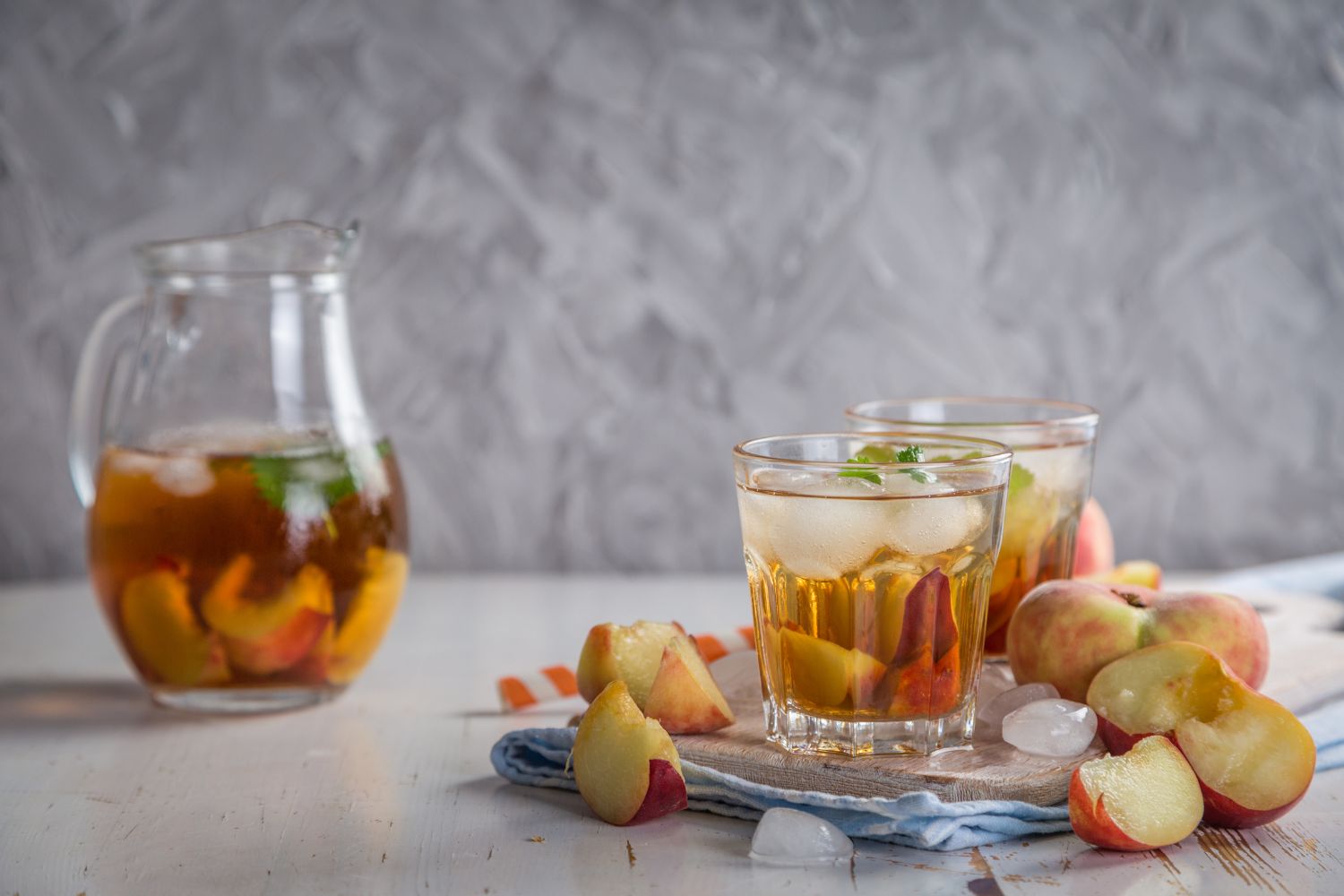 Gläser mit Eistee und Pfirsichstücken, Krug im Hintergrund. Dekor auf Tisch mit Eiswürfeln und ganzen Pfirsichen.
