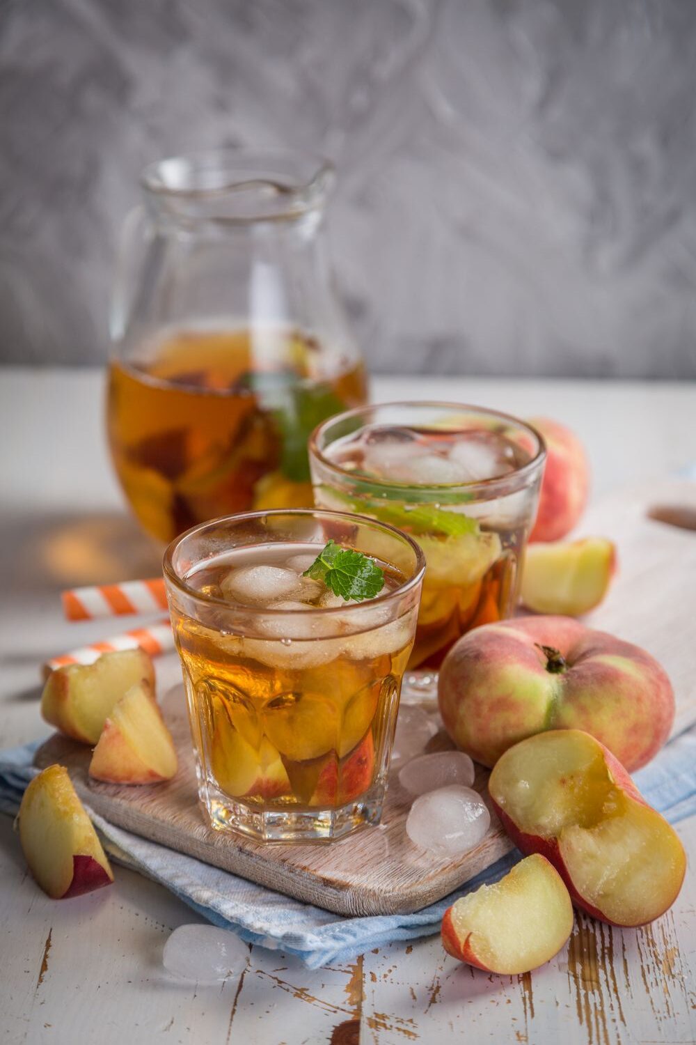Gläser mit eisgekühltem Pfirsich-Eistee und frischen Pfirsichstücken auf einem Holzbrett, umgeben von Eiswürfeln.
