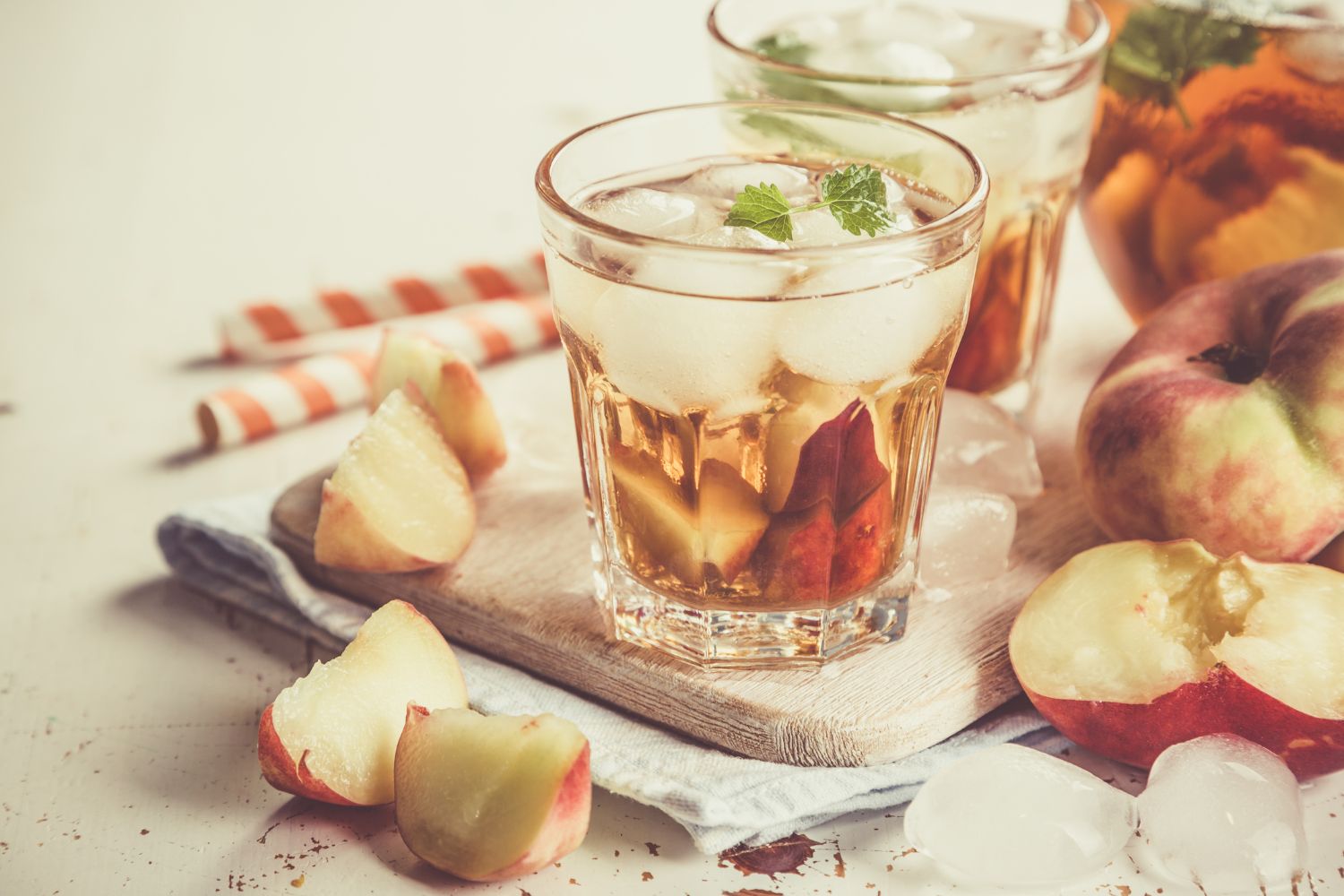 Glas Eistee mit Pfirsichstücken und Minze, daneben frische Pfirsiche und Eiswürfel auf einem Holzbrett.