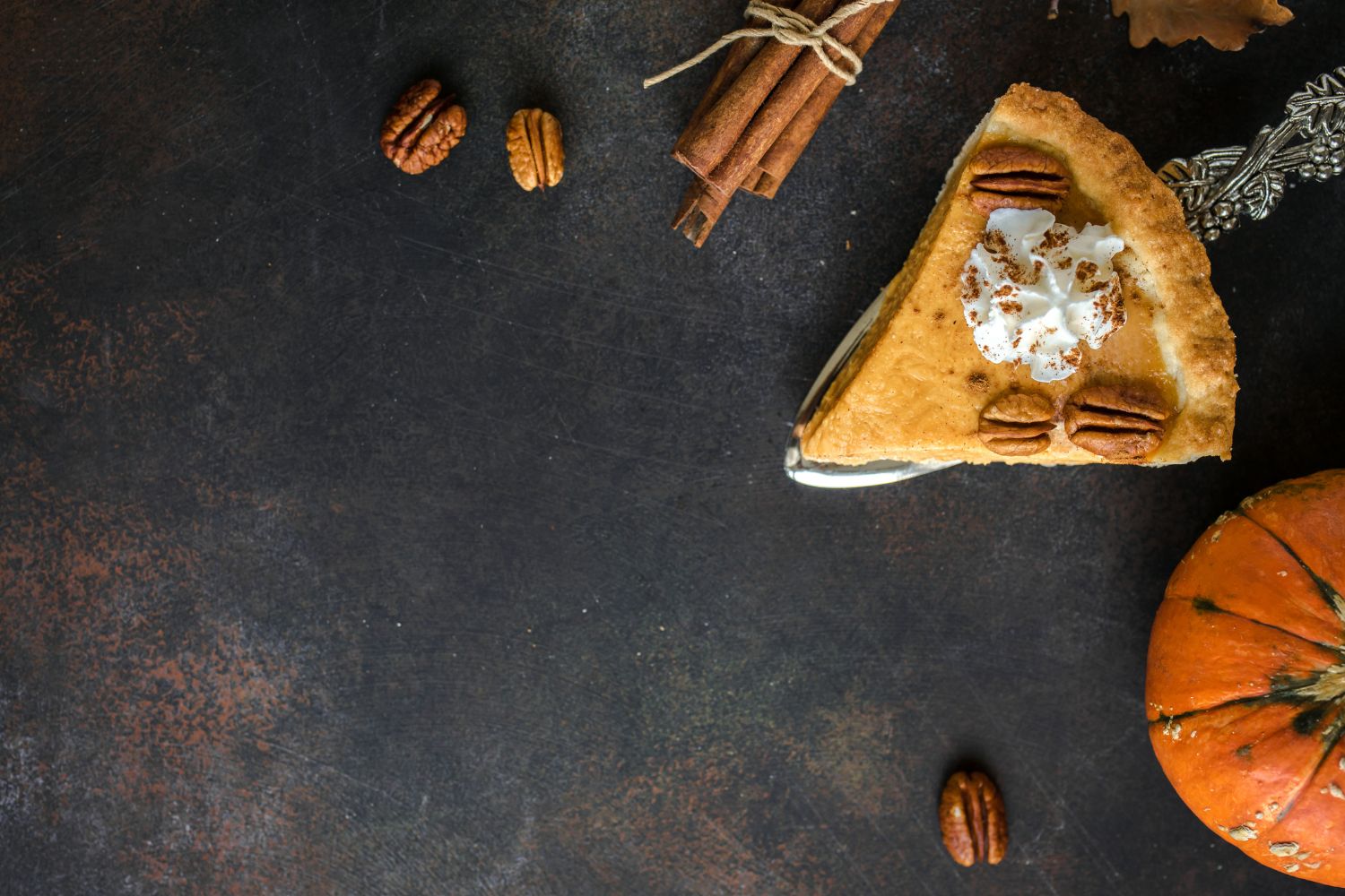 Kürbiskuchen Stück Kürbiskuchen mit Sahne und Pekannüssen auf einem dunklen Tisch, neben Zimtstangen und einem Kürbis.
