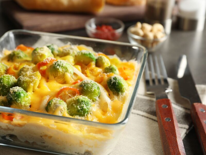 Auflauf in Glasform mit Rosenkohl und Käse überbacken auf Tisch mit Gabel und Messer.