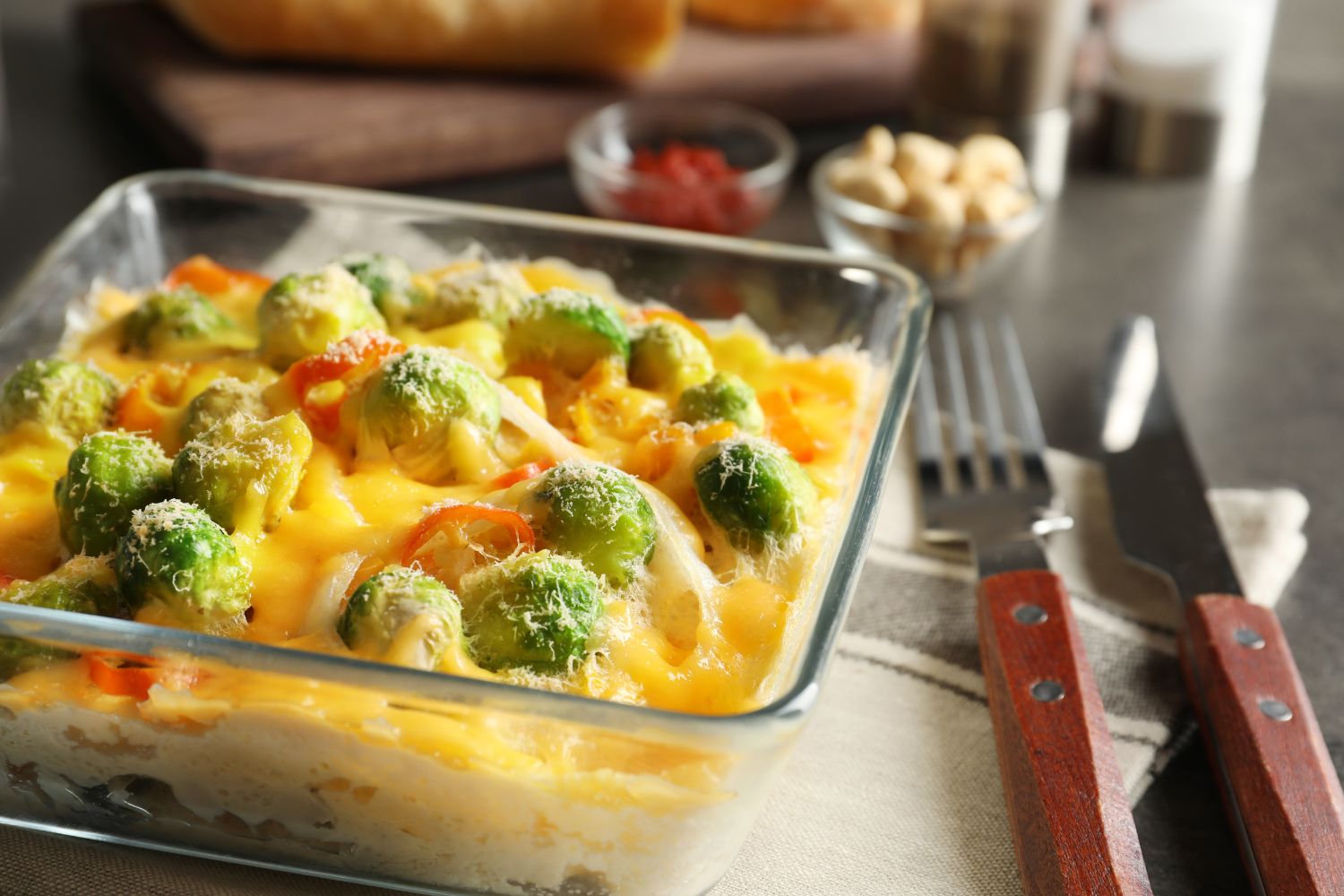 Auflauf in Glasform mit Rosenkohl und Käse überbacken auf Tisch mit Gabel und Messer.