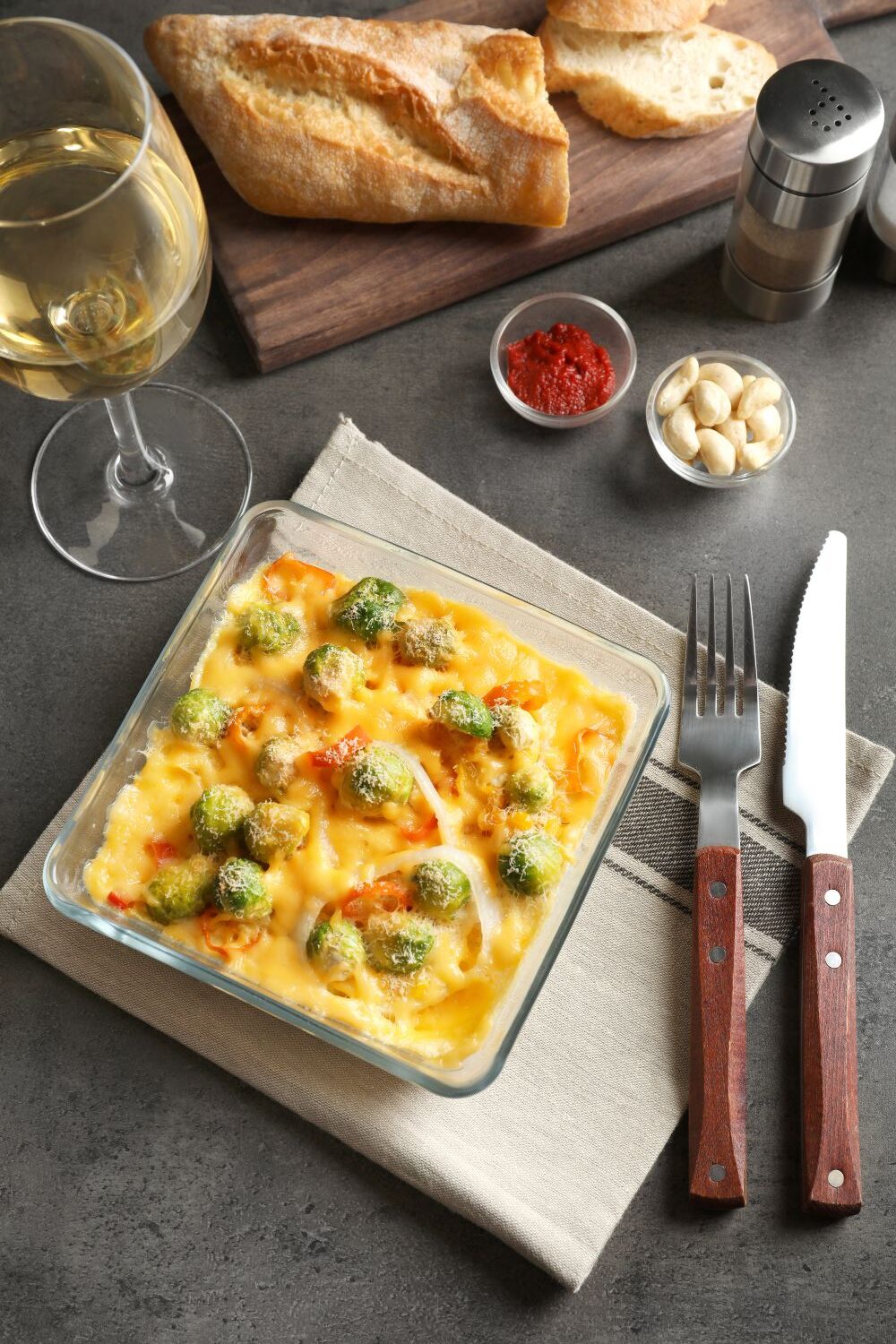 Auflauf in Glasform mit Rosenkohl und Käse überbacken auf Tisch mit Gabel und Messer.